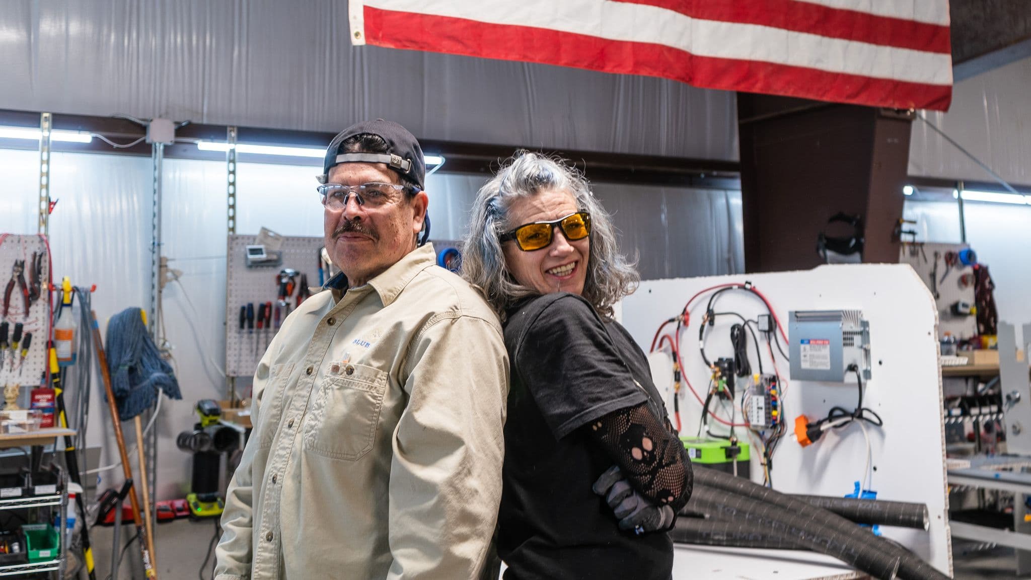 Two Escapod employees pose playfully in the factory, standing back-to-back with an American flag hanging behind them.