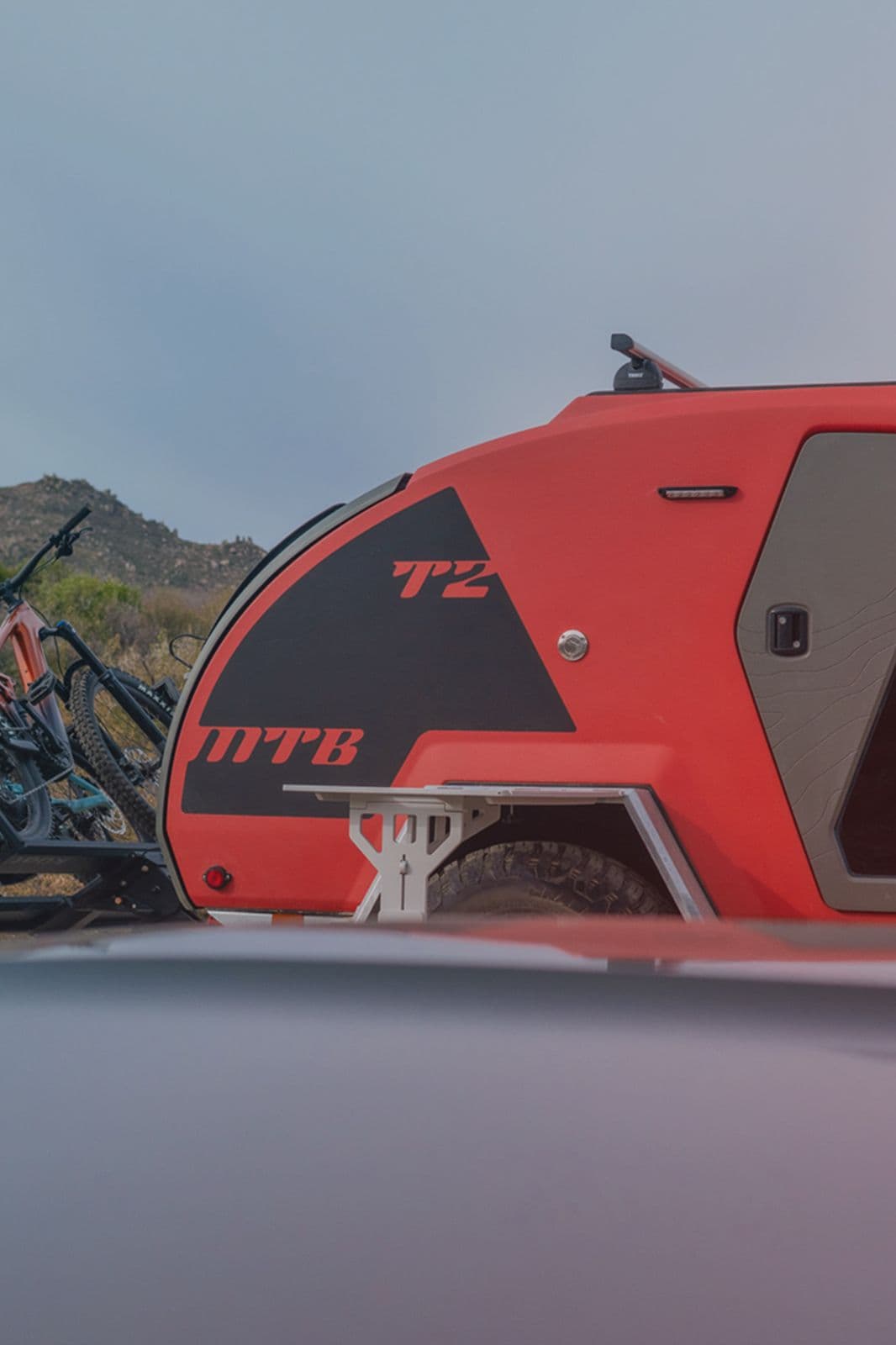 The rear of an orange TOPO2 MTB with bikes attached to the hitch, and the hood of a car blurred out in the foreground.