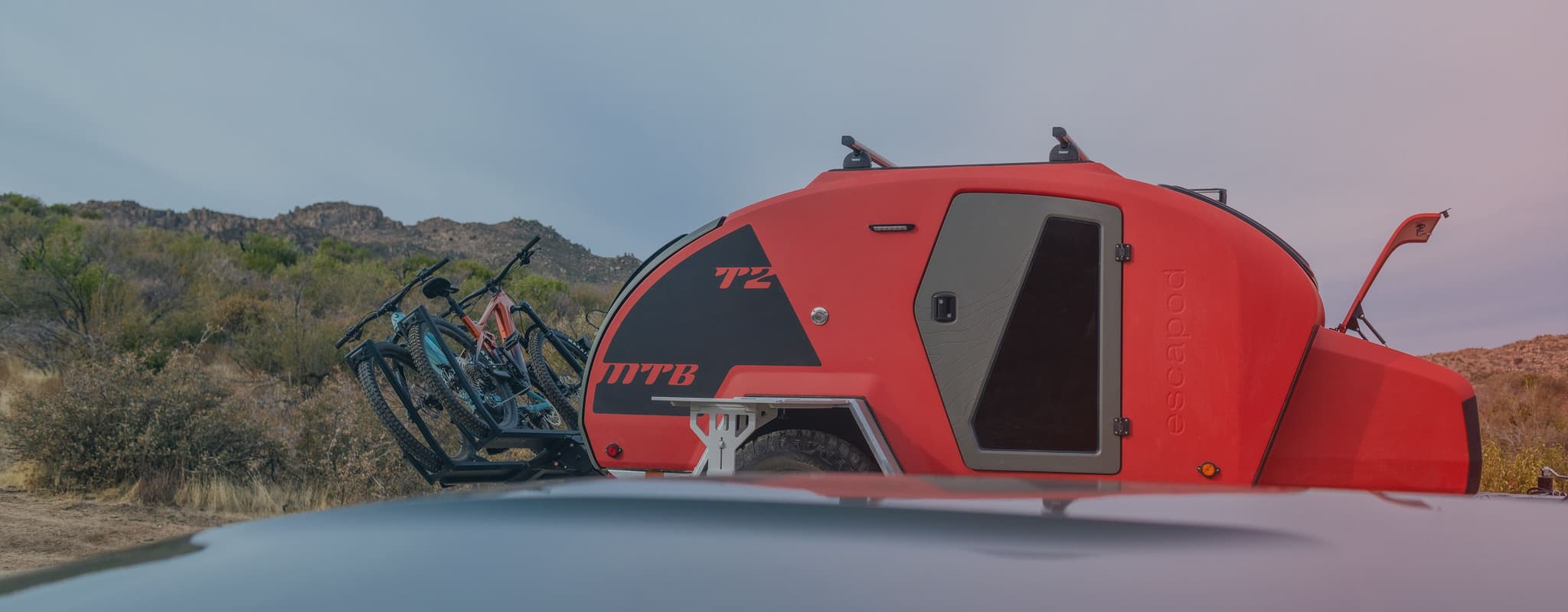 An orange TOPO2 MTB with the front tongue box opened and bikes attached to the hitch, and the hood of a car blurred out in the foreground. Desert bushes fill the background.