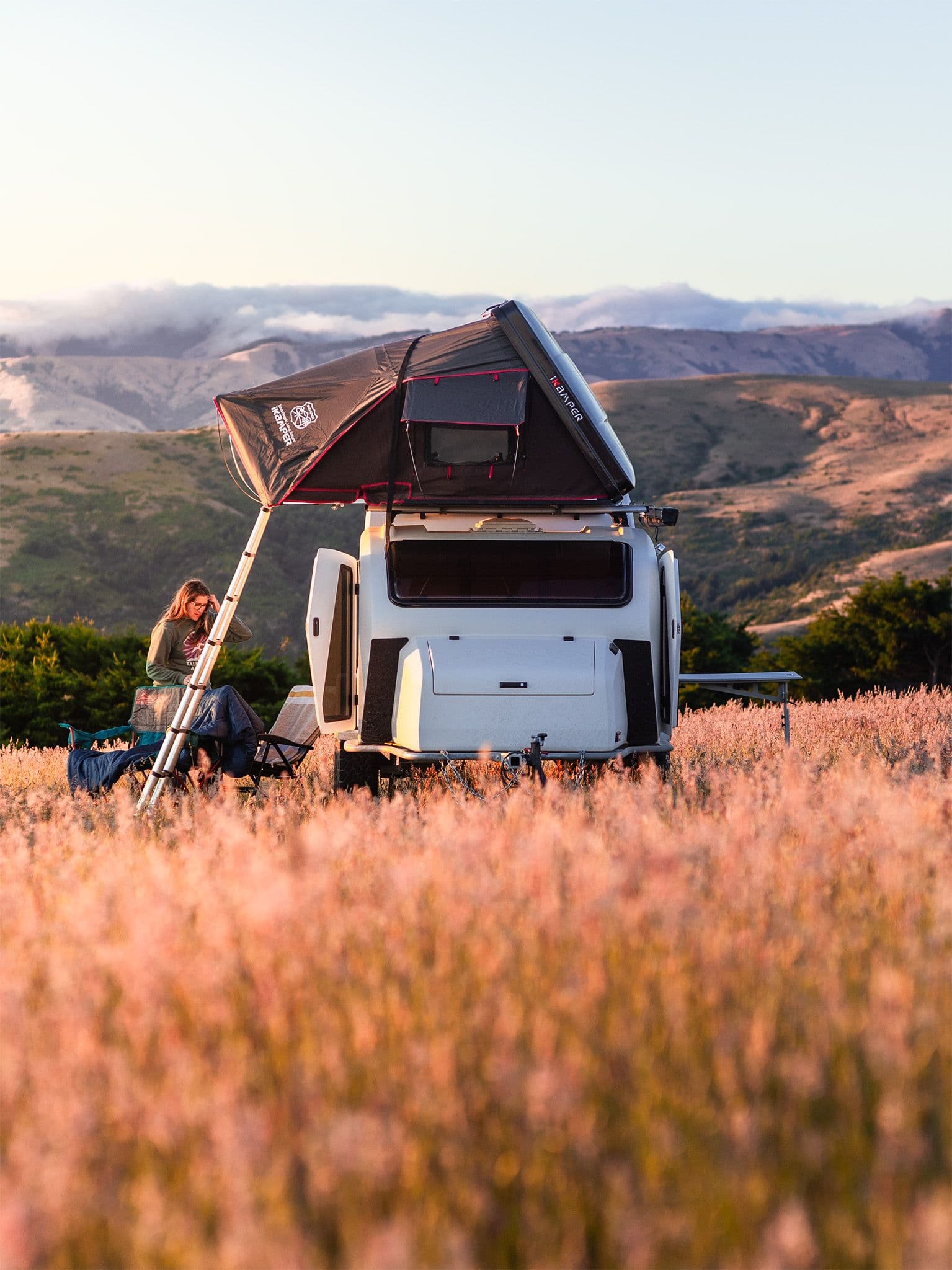 A white Escapod TOPO teardrop trailer with a rooftop tent set up in a golden field during sunset, surrounded by rolling hills.