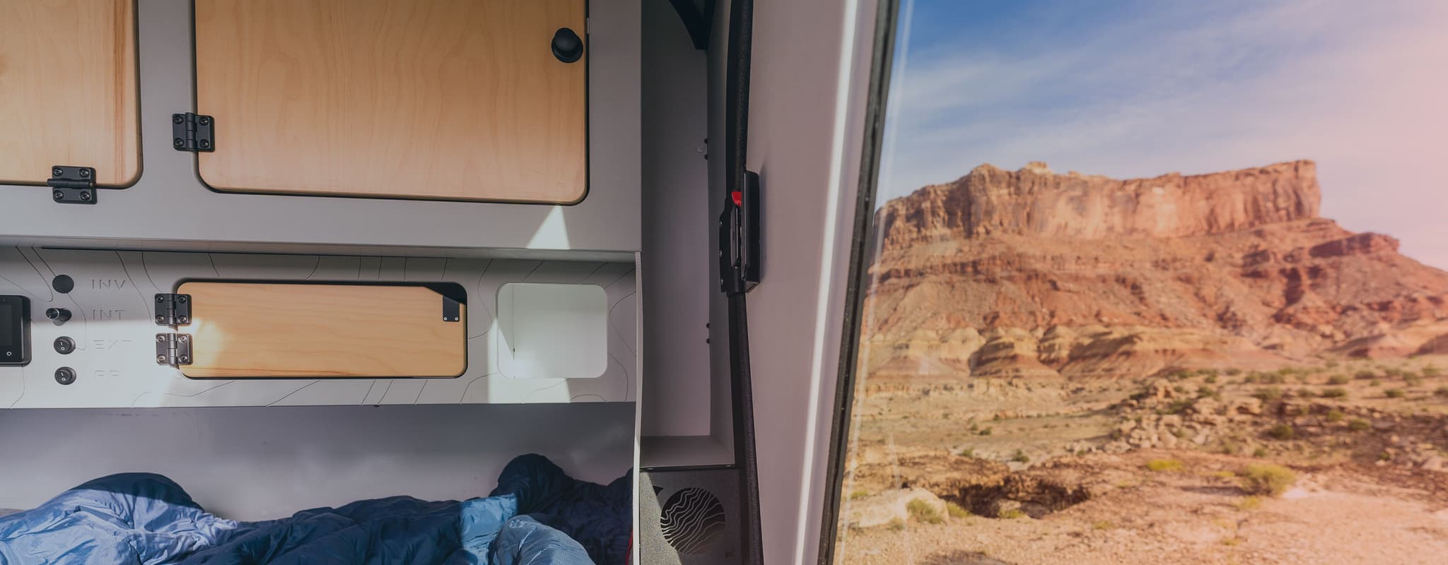 Interior of an Escapod TOPO2 trailer showcasing modern cabinetry and controls, with a view of a vast red rock desert landscape through the open door.