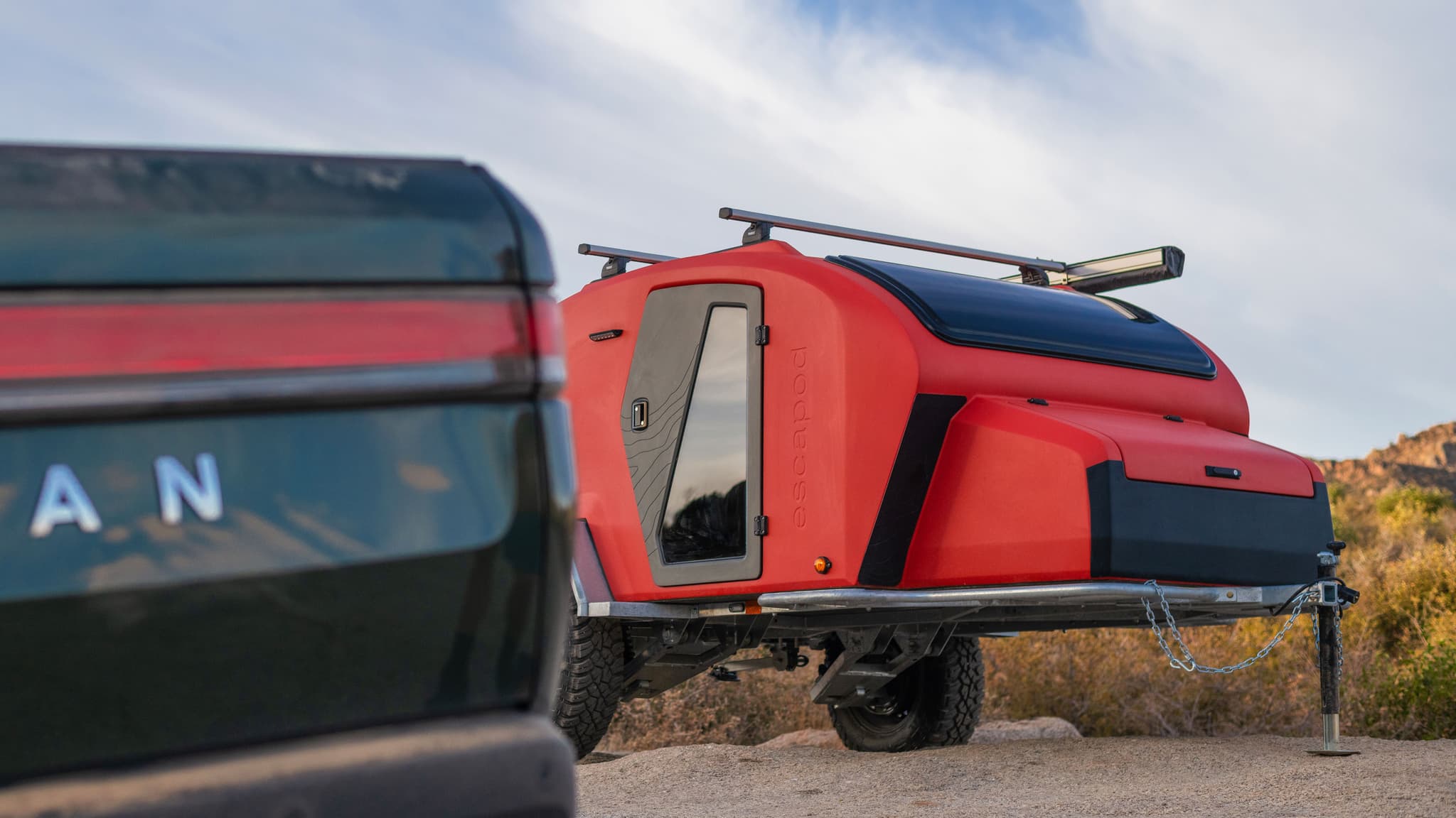 Rear three-quarter view of the red TOPO2 MTB trailer hitched to a green Rivian, with aerodynamic curves and rugged off-road tires.