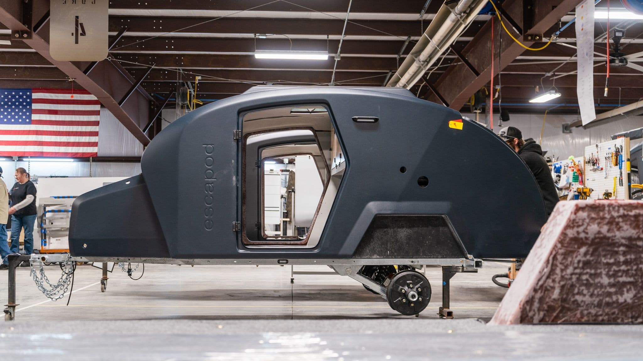 A close-up side view of an Escapod trailer body under assembly, with an open door revealing the unfinished interior and team members working in the background.