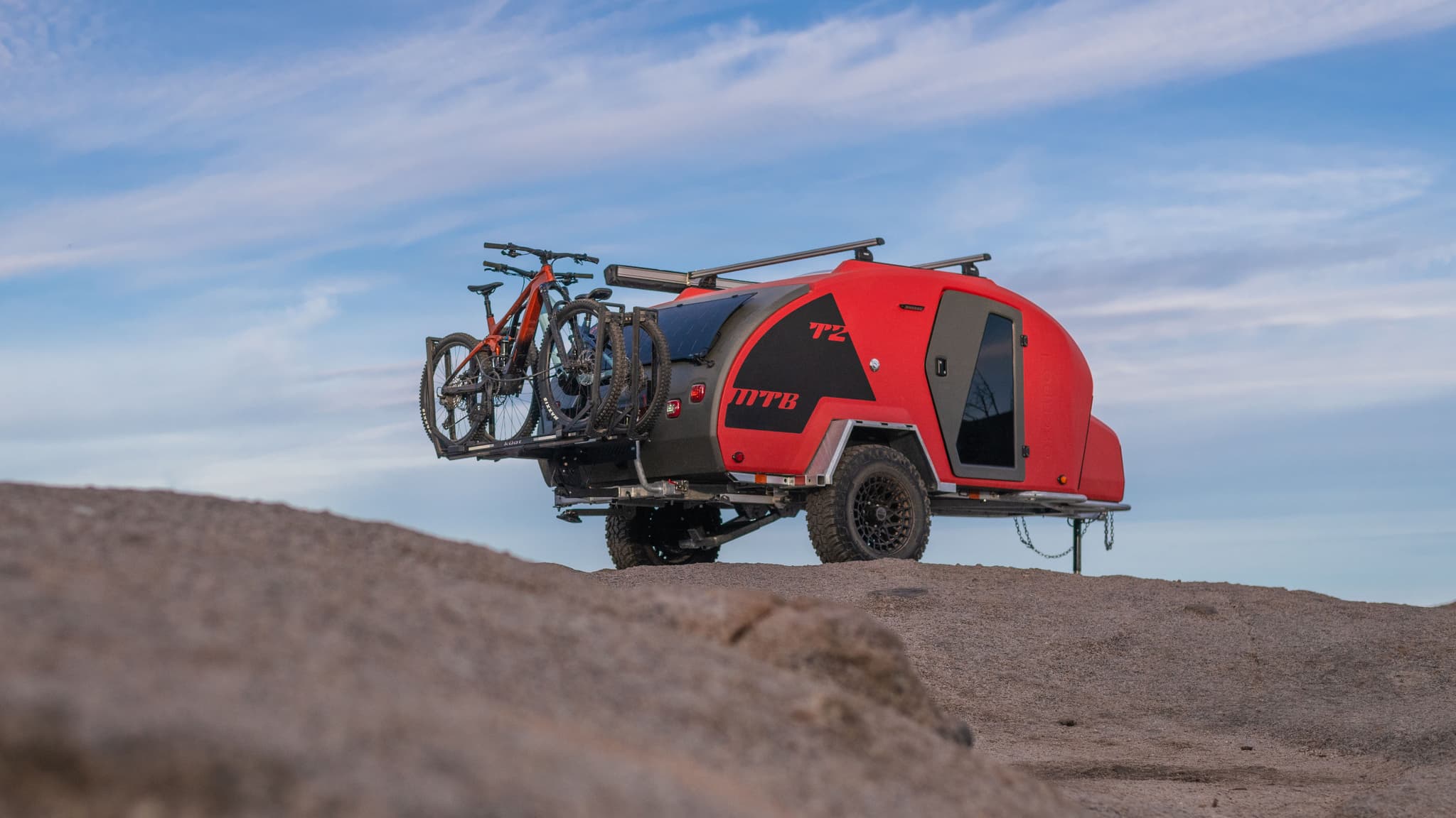 Angled rear view of the TOPO2 MTB trailer with bikes mounted on the back, parked on a rocky slope under a blue sky with clouds.