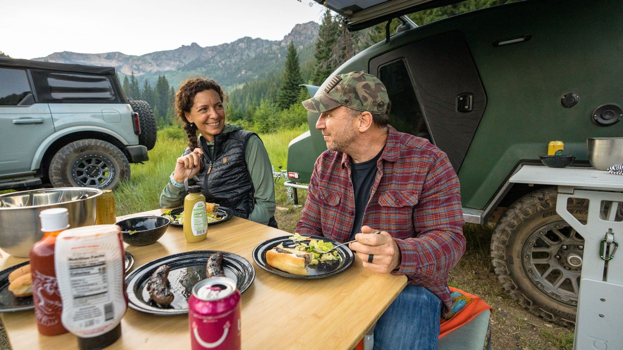 Couple enjoying a campfire meal outside their Escapod trailer with a Ford Bronco nearby, surrounded by lush green mountains at sunset.