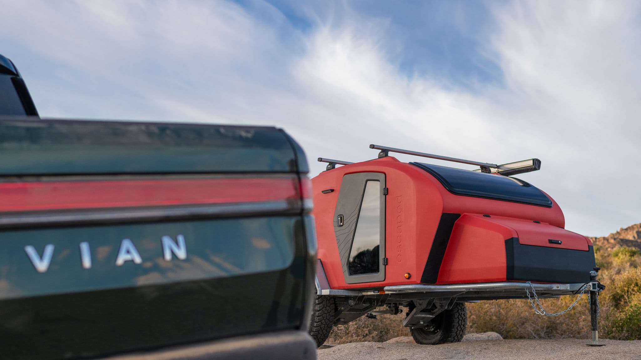 A orange Escapod TOPO2 MTB trailer parked beside a green Rivian truck in a desert landscape, highlighting its compact, off-road design.