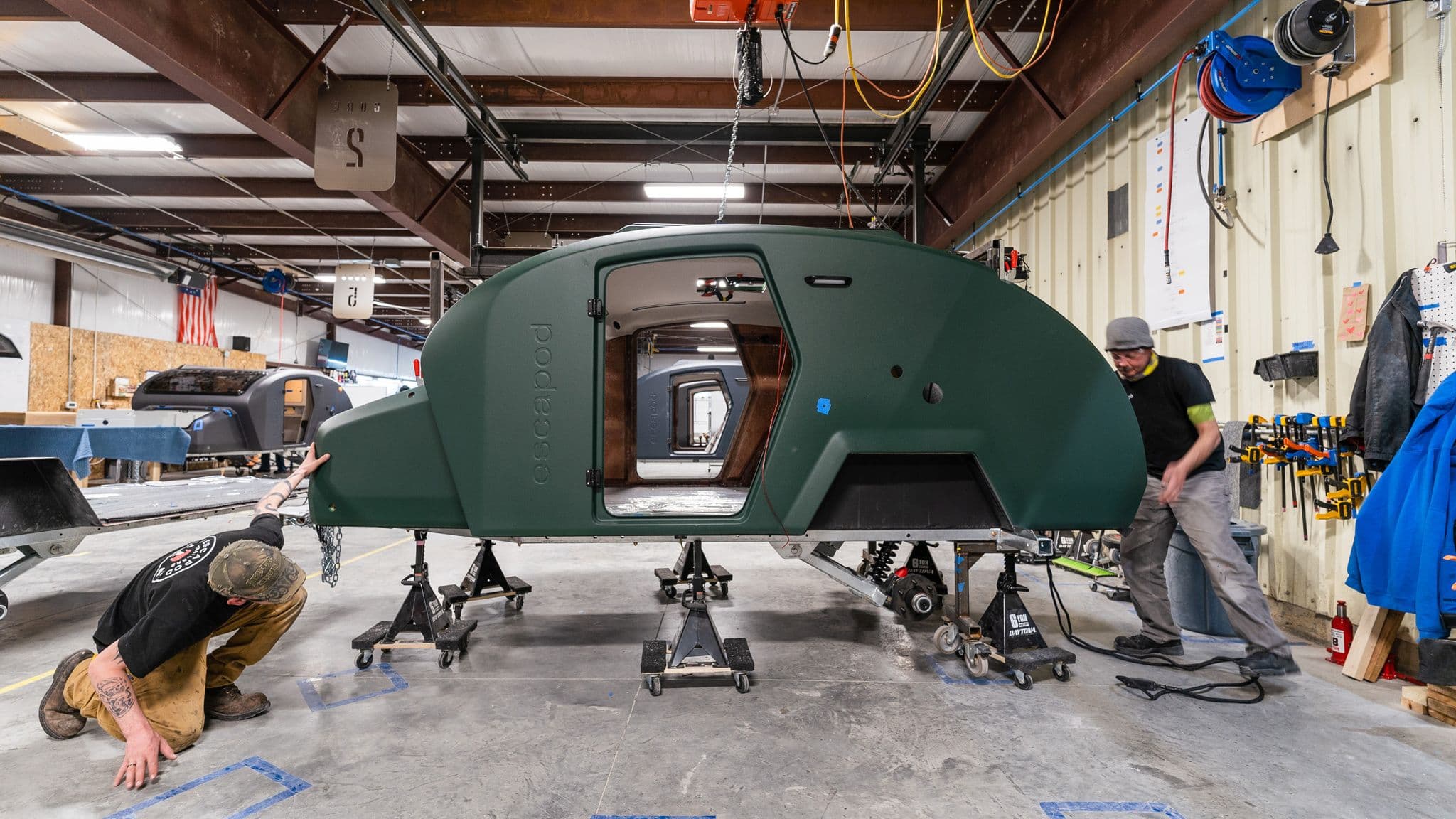Two workers positioning a green Escapod TOPO2 trailer on an assembly stand, aligning it for further installation and finishing.
