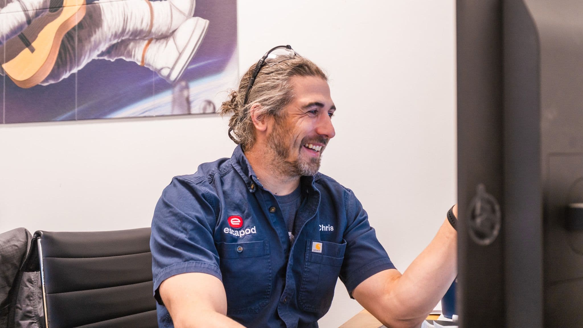 An Escapod employee named Chris, wearing a navy work shirt with an embroidered logo, smiles at his desk while working on a computer in an office setting.