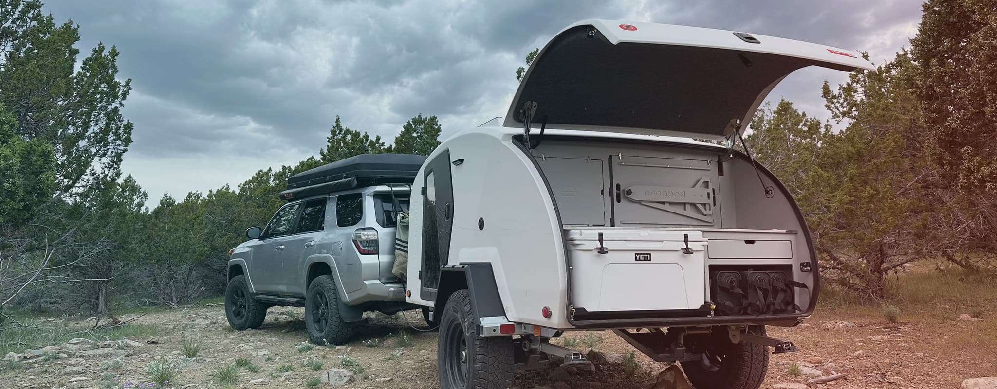 A white Escapod TOPO2 Nomad with its galley open, showing the kitchen and white YETI cooler, photographed on rocky terrain with a cloudy sky, being towed by a Toyota 4Runner