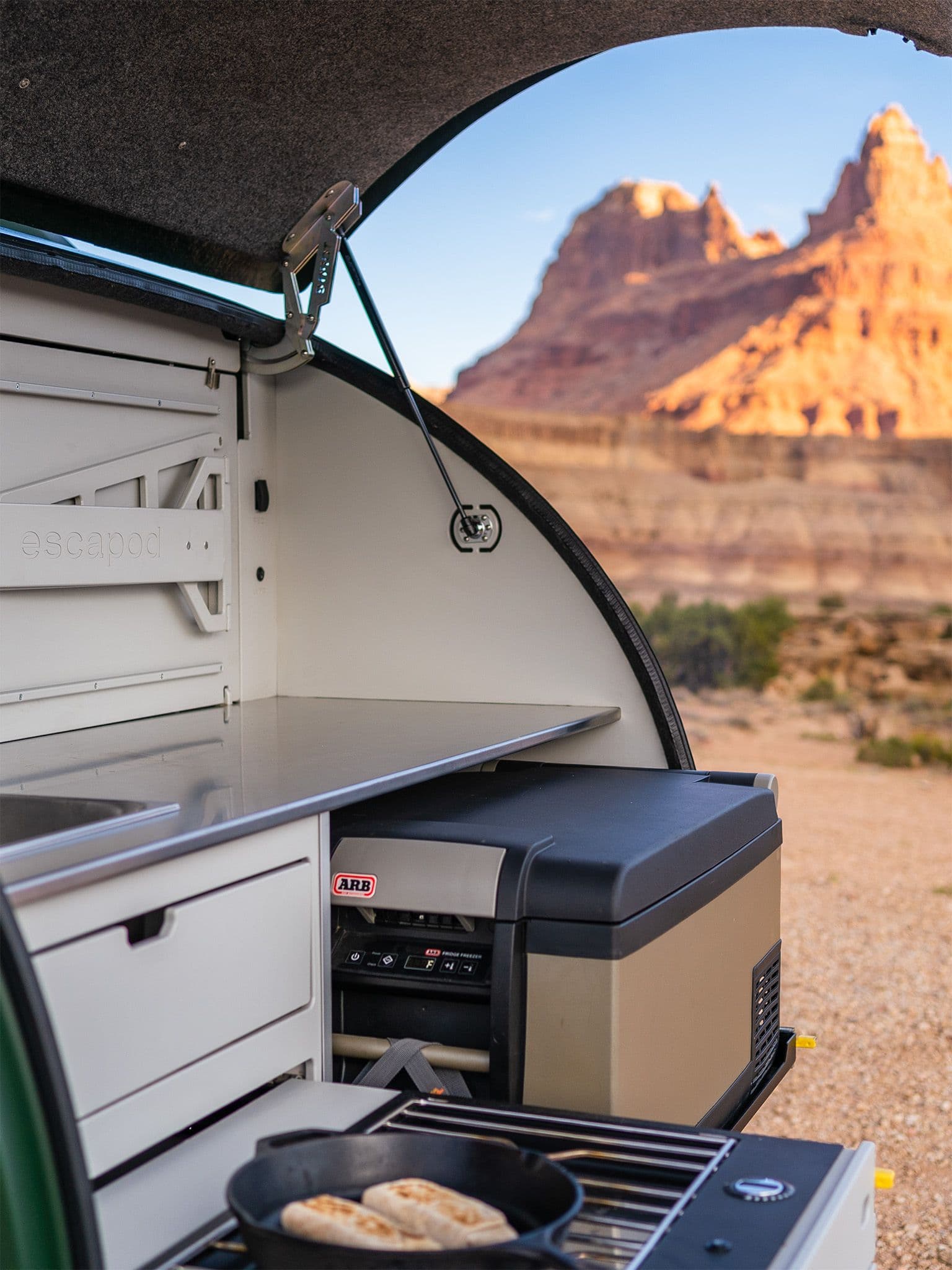 The galley of a TOPO2 at sunset in Utah, showing a cast iron skillet on the stove with two burritos, an ARB fridge halfway pulled out, and the stainless steel countertop.