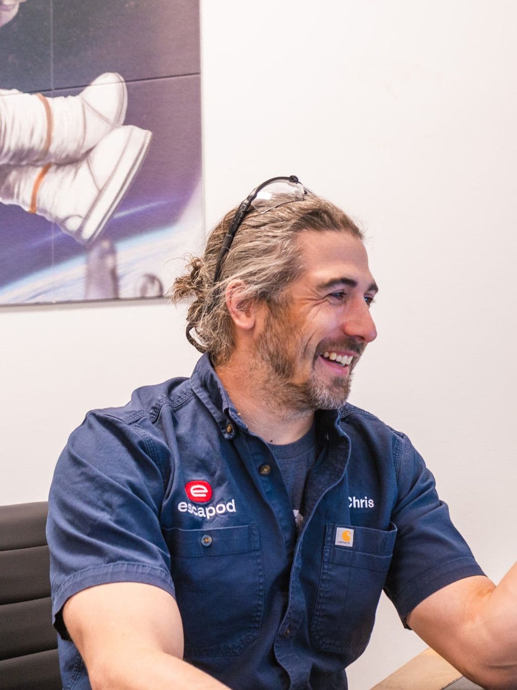 An Escapod employee named Chris, wearing a navy work shirt with an embroidered logo, smiles at his desk while working on a computer in an office setting.