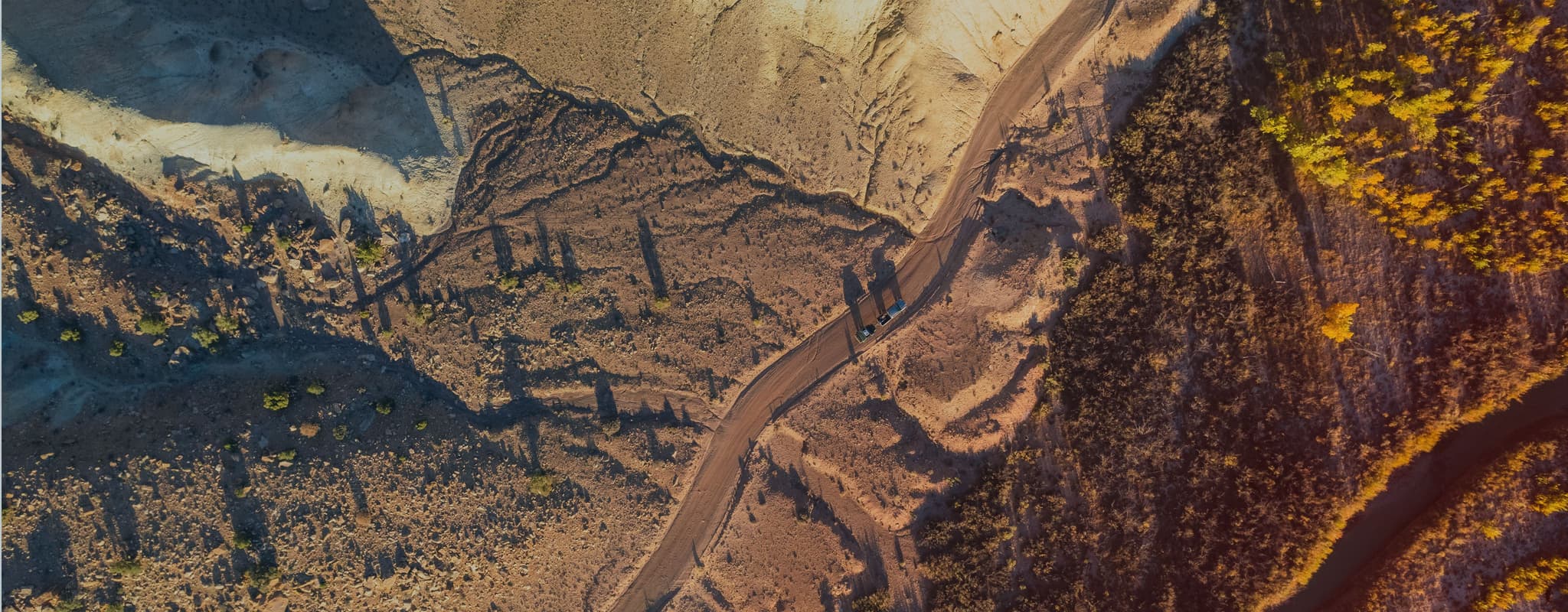Aerial shot of two off-road trailers and vehicles driving along a winding dirt road through a rugged desert landscape at sunset.