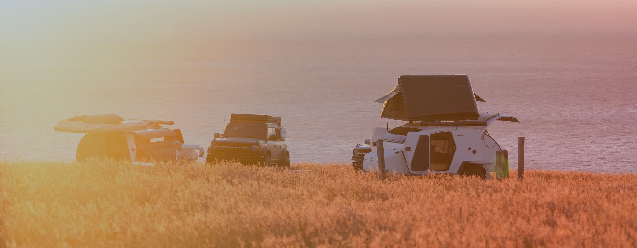 Two Escapod TOPO2 trailers set up for camping on a scenic coastal bluff, bathed in warm golden light as the sun sets over the ocean horizon.