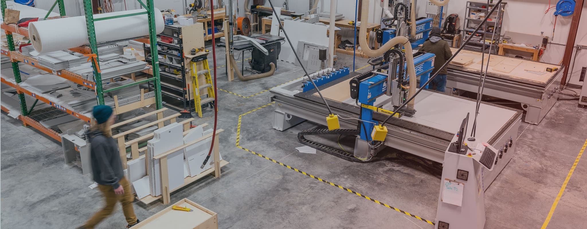 A spacious manufacturing facility with CNC machines cutting materials, organized storage racks, and workers moving through the space.