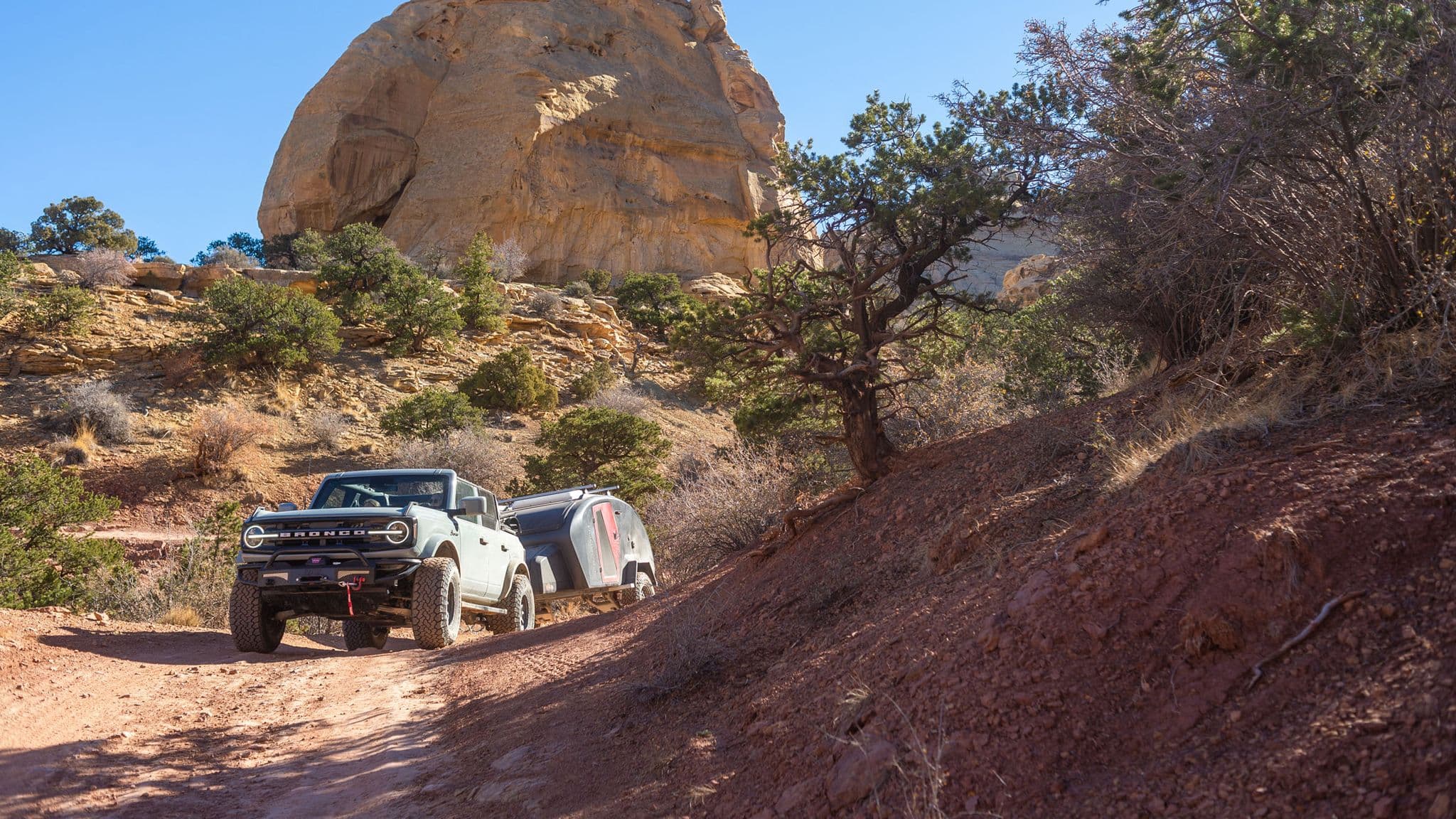 Ford Bronco towing an Escapod trailer on a rugged dirt road through a canyon landscape.
