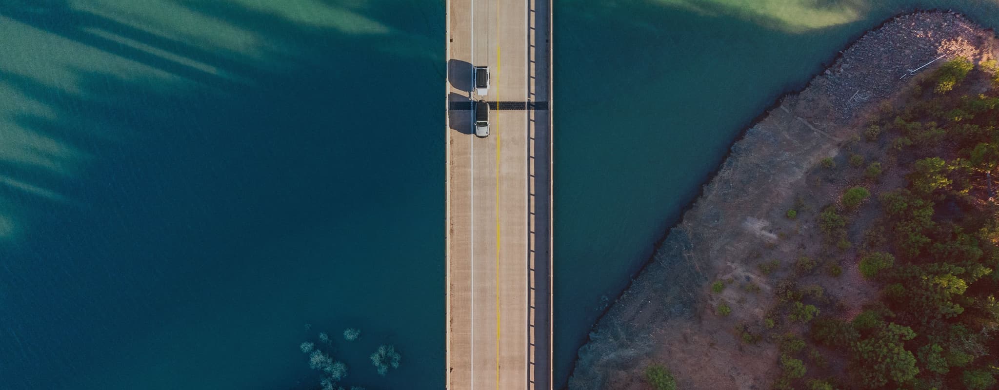 A drone view of an overland vehicle towing an Escapod TOPO2 trailer across a long bridge, surrounded by deep blue water and rugged shoreline.